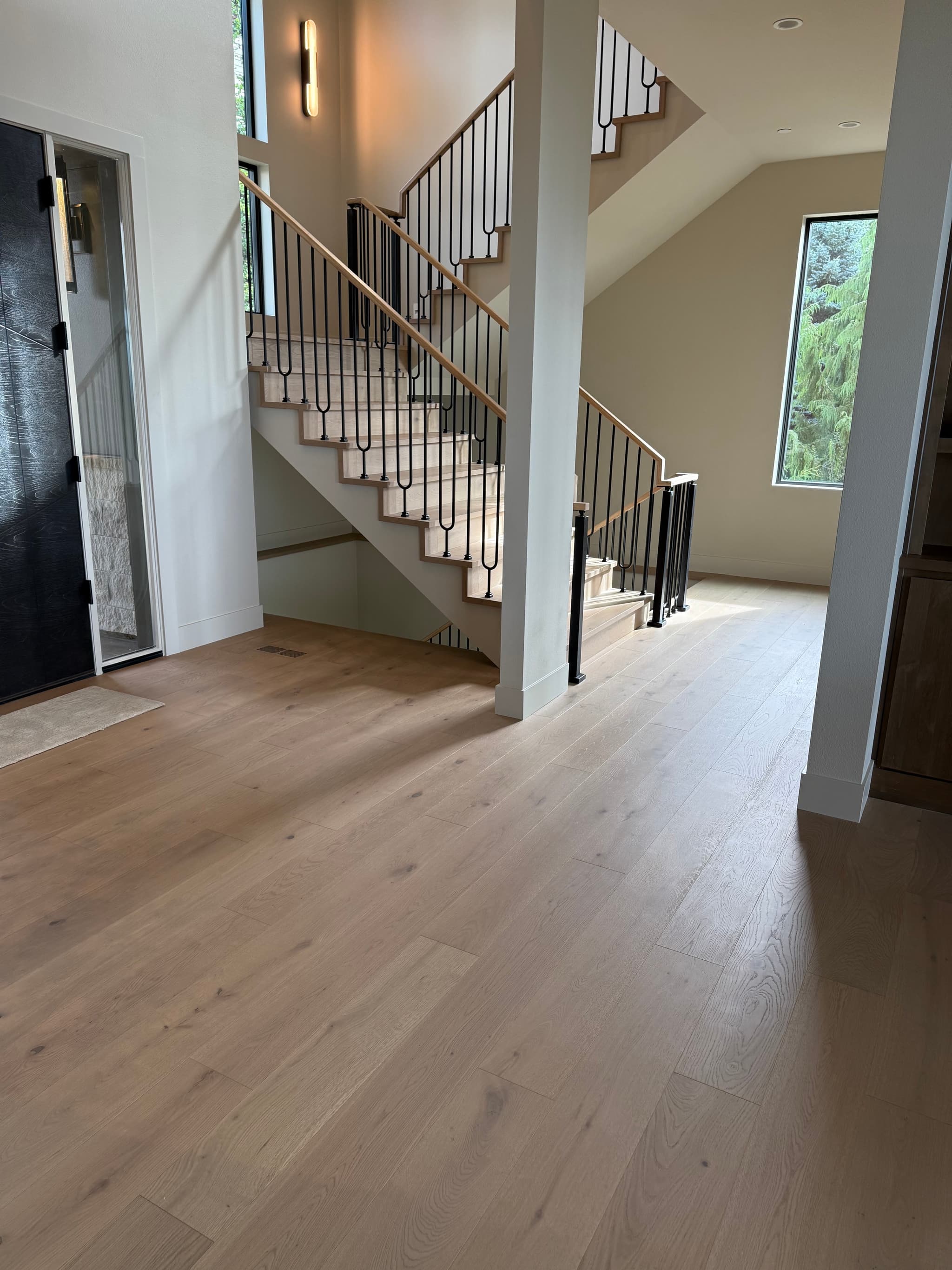 Engineered white oak hardwood flooring in a two-story entryway with a matching oak staircase, black iron balusters, and a black front door — Skyview Flooring project in Vancouver, WA