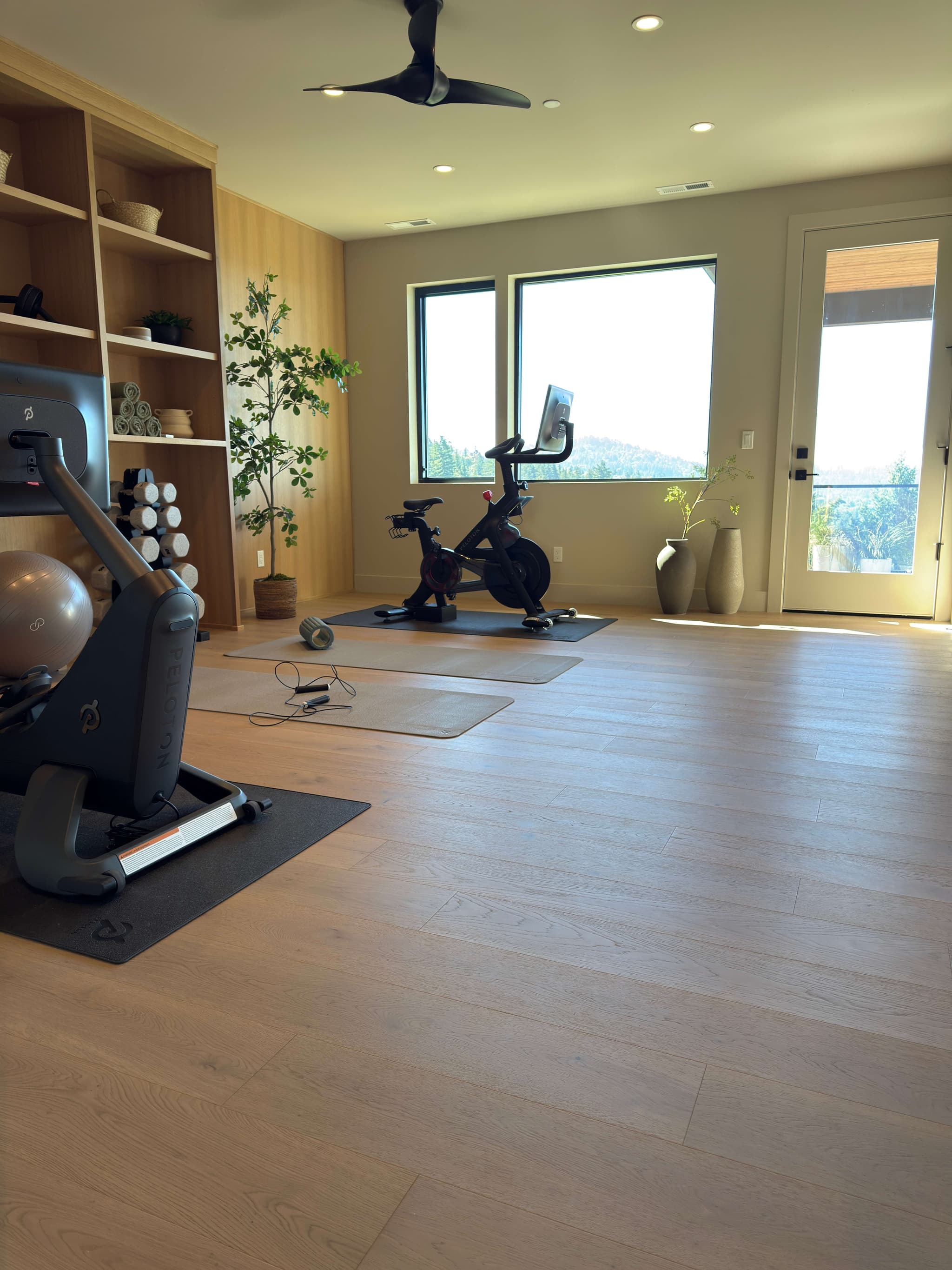 Wide-plank engineered white oak hardwood flooring in a home gym with a Peloton bike and tread, oak built-in shelving, dumbbell rack, and forest views through black-framed windows — Skyview Flooring project in Bethany, OR