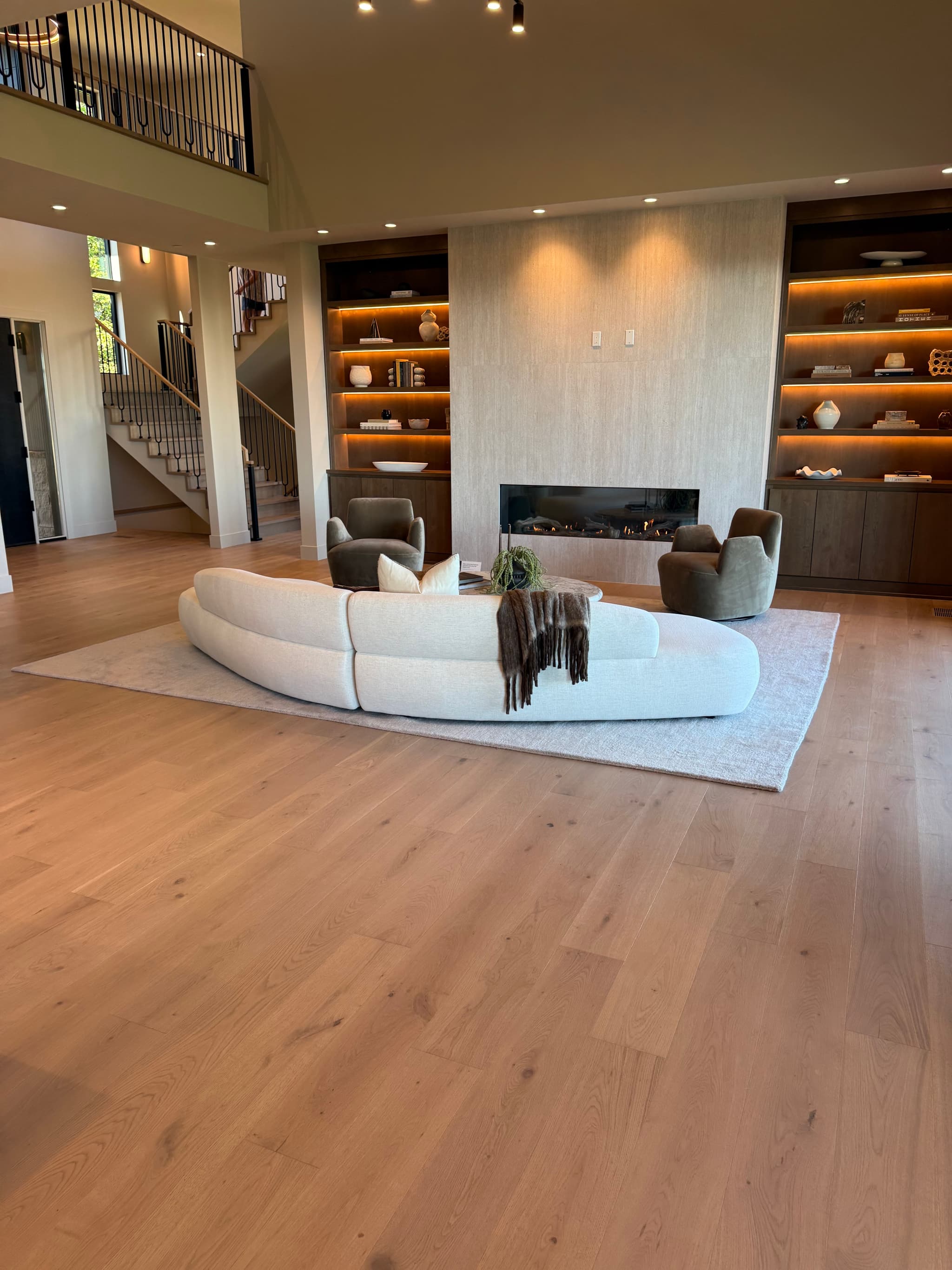 Wide-plank engineered white oak hardwood flooring in a two-story great room with a curved cream sectional, linear gas fireplace, and lit walnut built-in shelving — Skyview Flooring project in Bethany, OR