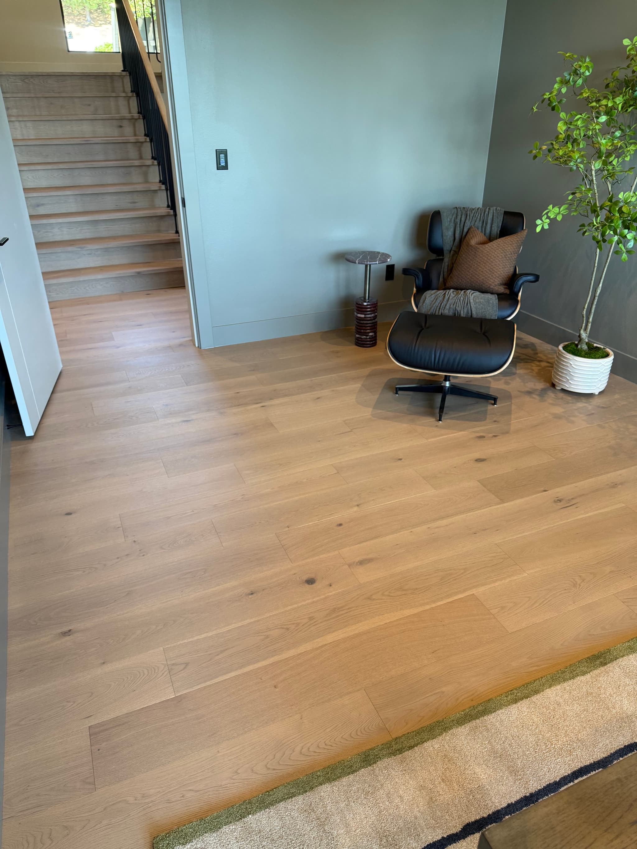 Wide-plank engineered white oak flooring installed in a home office with matching oak stair treads, a black leather lounge chair, and a carpet transition — Skyview Flooring project in Bethany, OR
