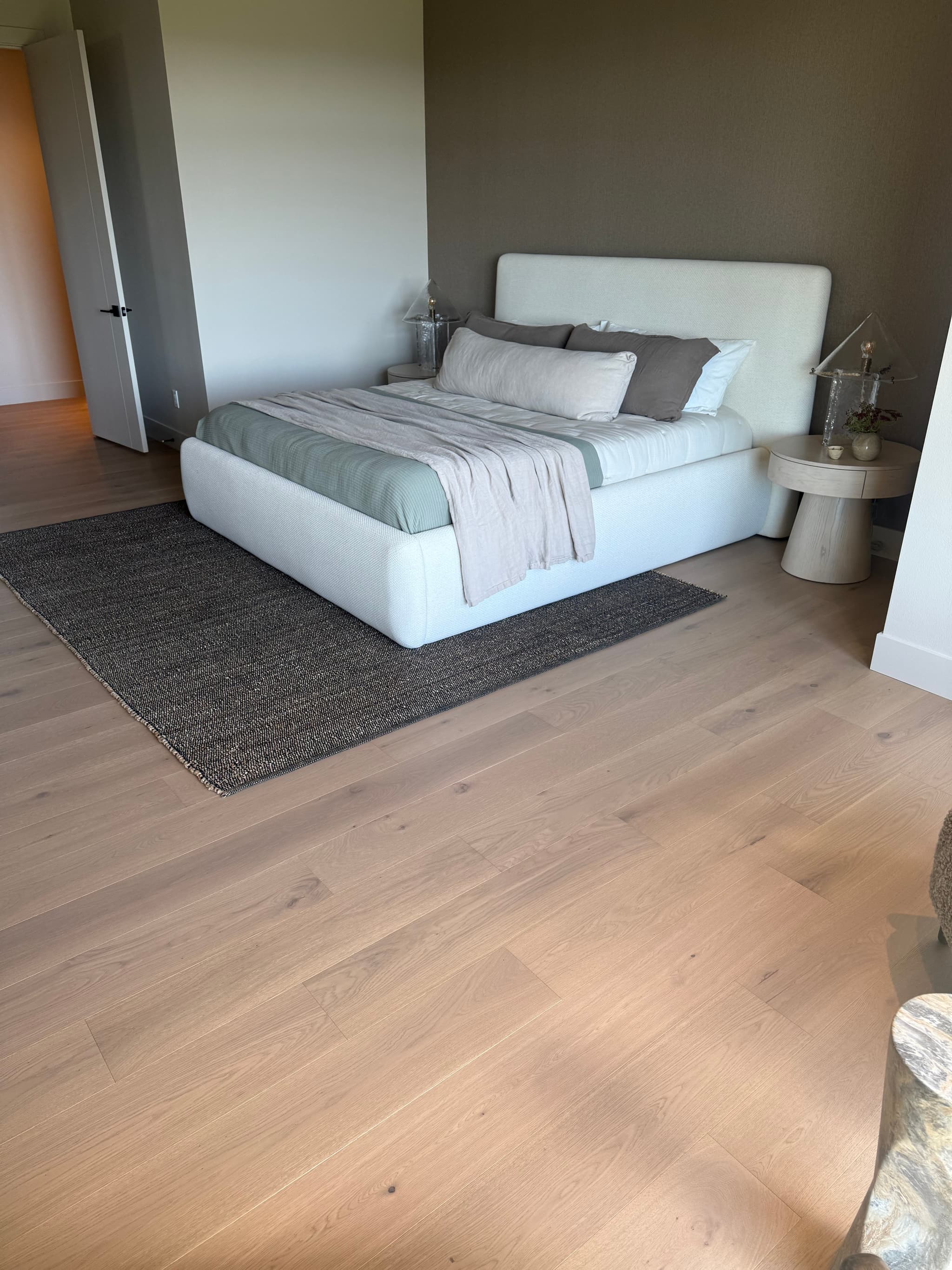 Light engineered white oak hardwood flooring in a primary bedroom with an upholstered white platform bed, charcoal woven rug, and round oak nightstand — Skyview Flooring project in Bethany, OR
