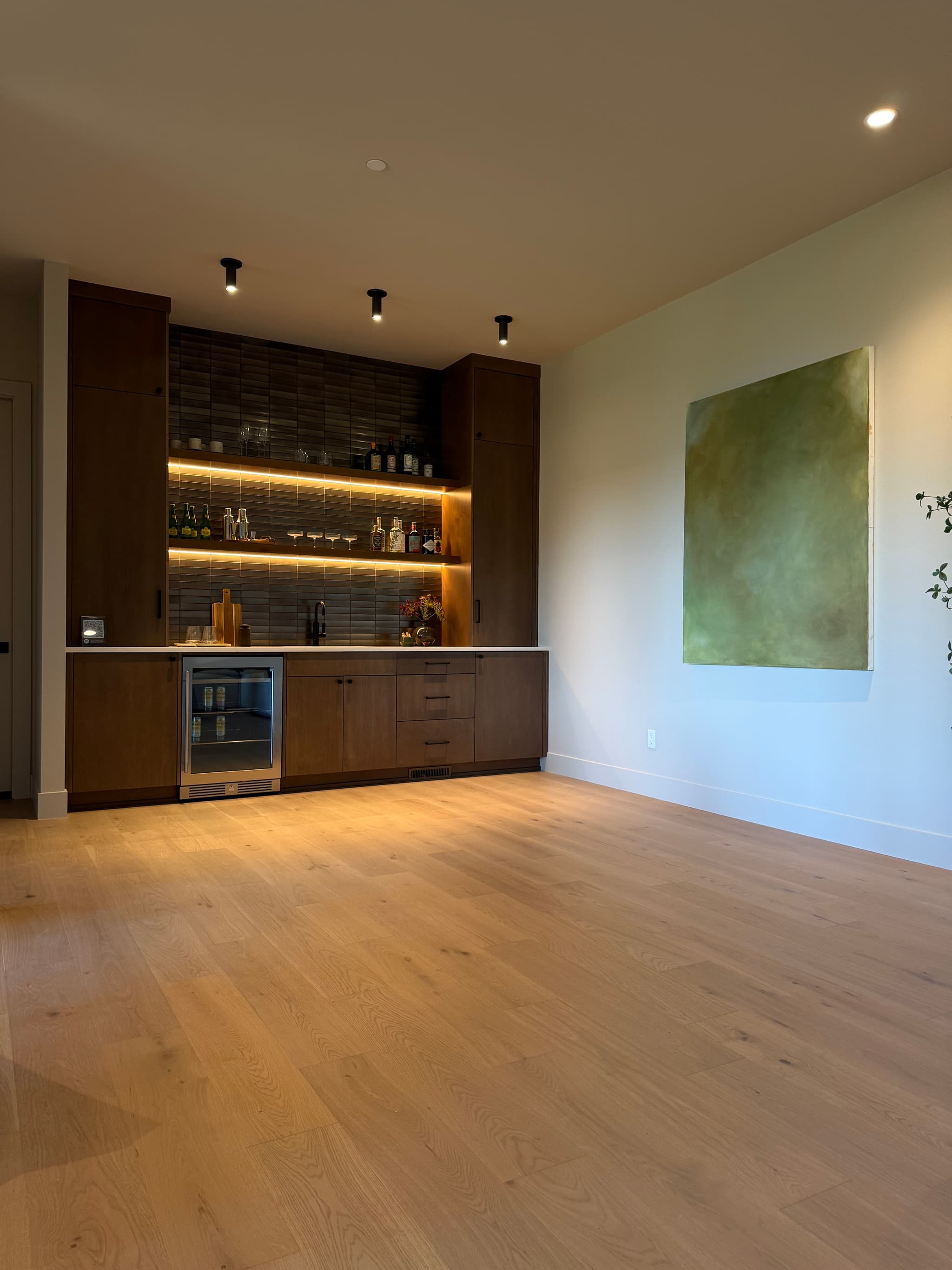 Engineered white oak hardwood flooring in a lower-level lounge with a walnut wet bar, LED-lit shelving, beverage fridge, and large green canvas artwork — Skyview Flooring project in Bethany, OR